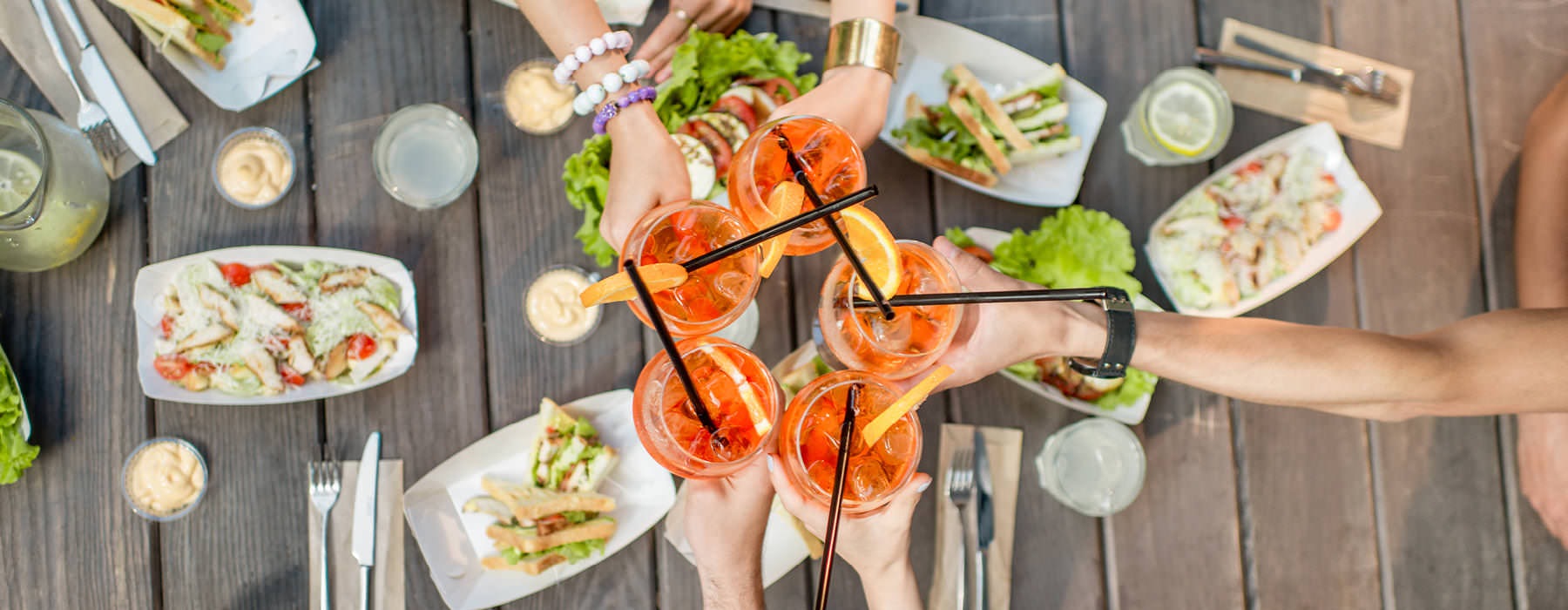 Group of people toasting glass cups of iced, orange beverages with straws and orange slices in them as they sit around a picnic table of food